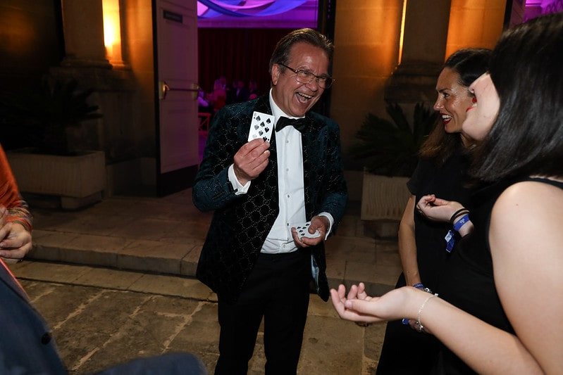The magician holding up a chosen card, guests laughing beside him at an evening reception