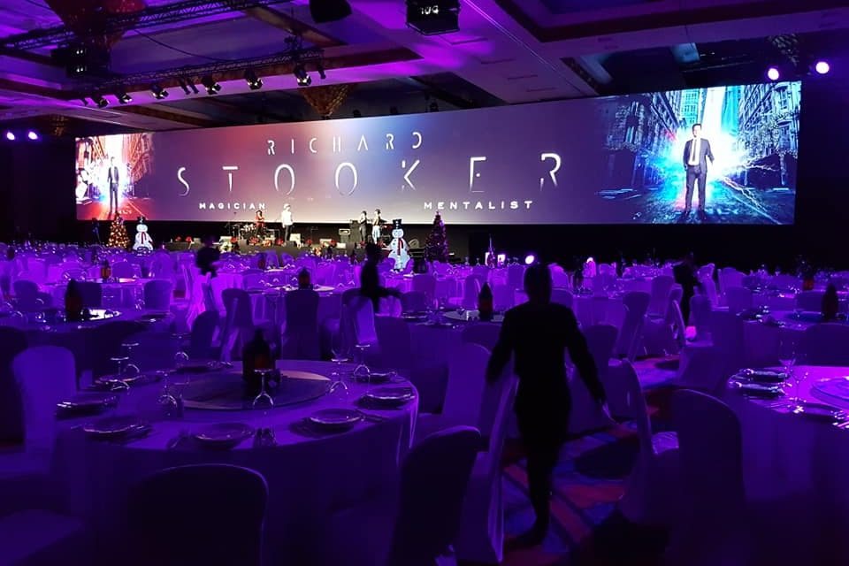 A banquet room set for dinner with a wide stage screen carrying the show titles