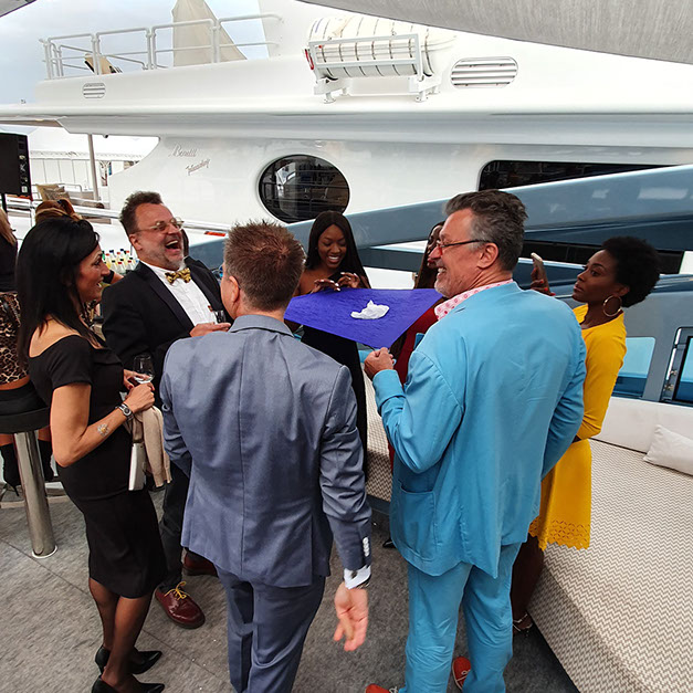 Guests standing on a yacht's aft deck in daylight, watching something held up in front of them